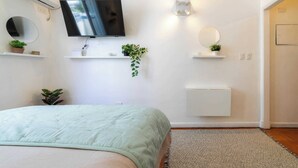 Ein Schlafzimmer mit einem Bett, einem an der Wand montierten Fernseher, einer Toilette und einem kleinen Regal mit einer Pflanze.