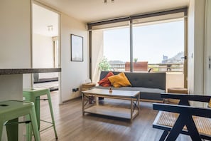 A modern living room with a sofa, coffee table, and green bar stools. A large window offers a view of buildings and a balcony. - Live the authentic atmosphere of Barrio Italia (Ñuñoa)