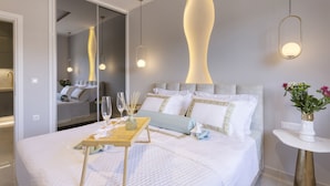 A modern hotel room with a neatly made bed, a bedside table with a vase of flowers, and a large mirror reflecting the room. - Gold Apartment (Nea Smirni)