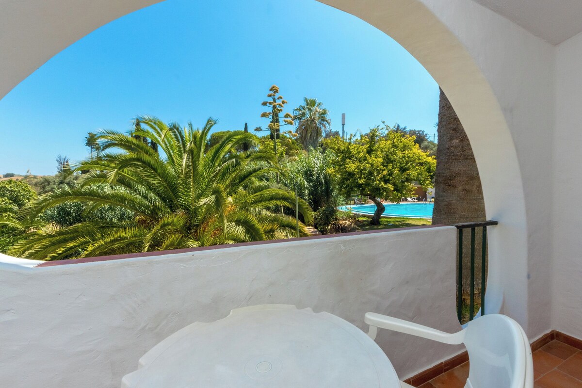 A balcony with a view of a swimming pool, palm trees, and a clear blue sky.
