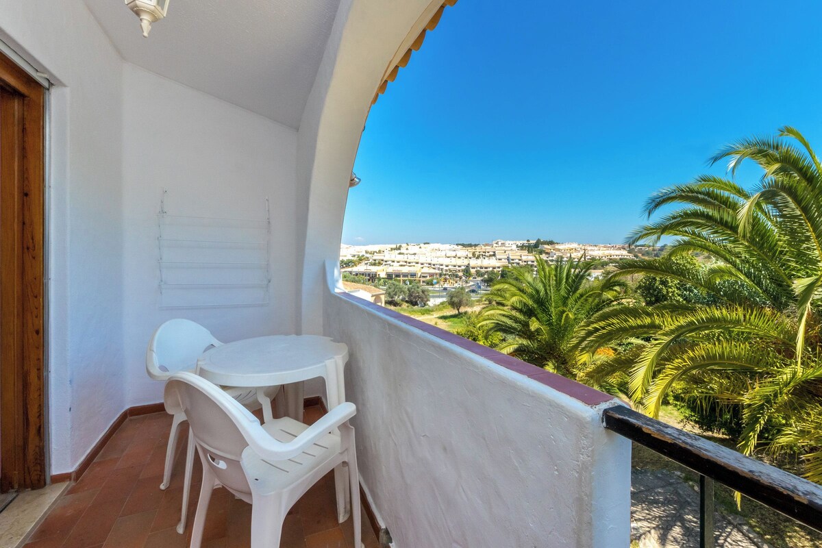 A balcony with a white table and chairs, a view of a cityscape, and palm trees.