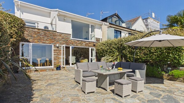 Une maison moderne avec une façade en pierre, de grandes fenêtres et une terrasse aménagée avec du mobilier d’extérieur.