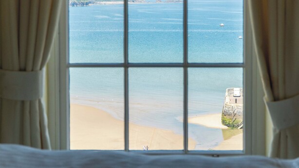 Une vue à travers une fenêtre donnant sur la mer, une plage de sable et une construction en pierre.