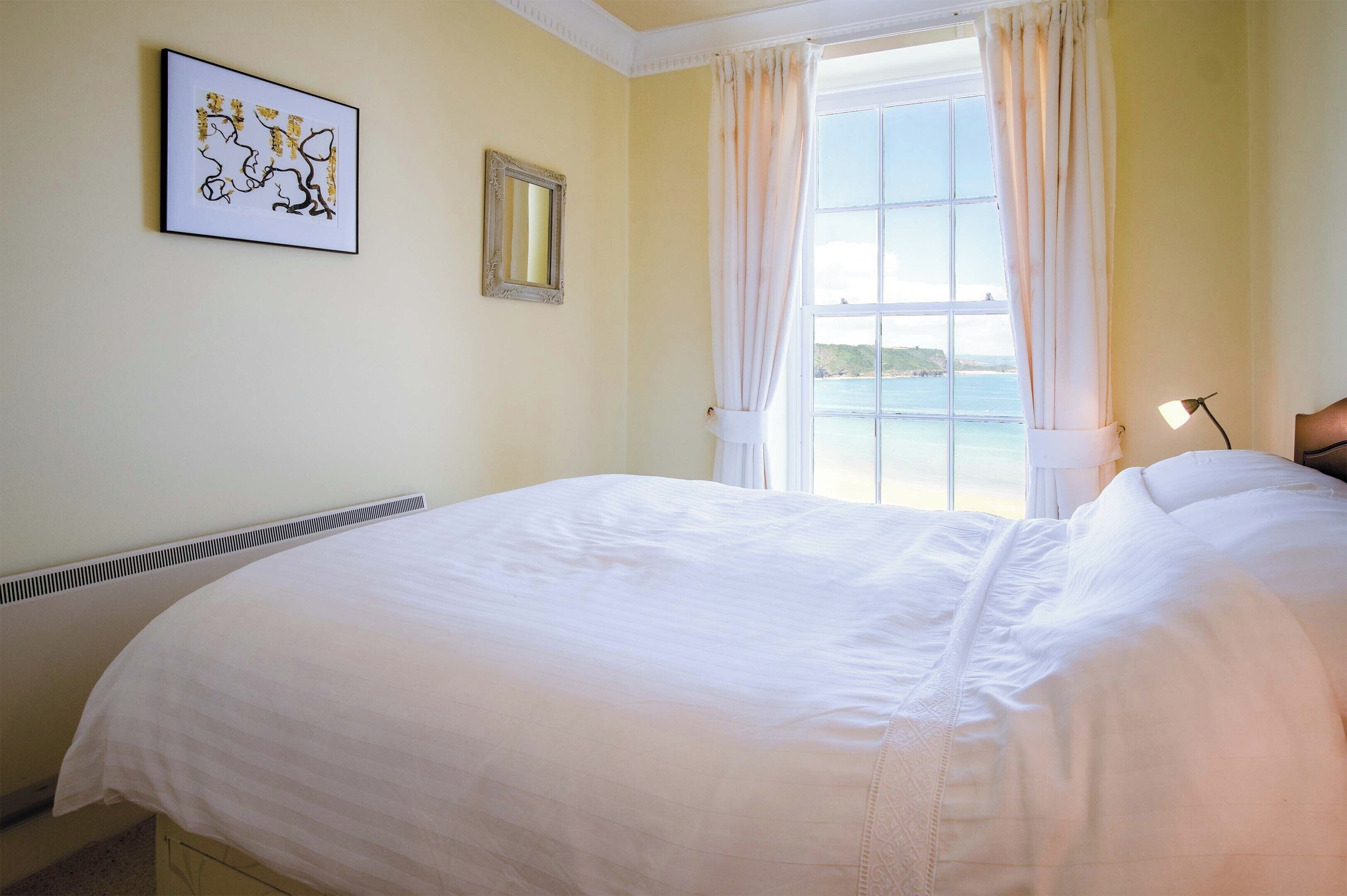 A neatly made bed in a room with a large window, a framed picture on the wall, and a radiator.