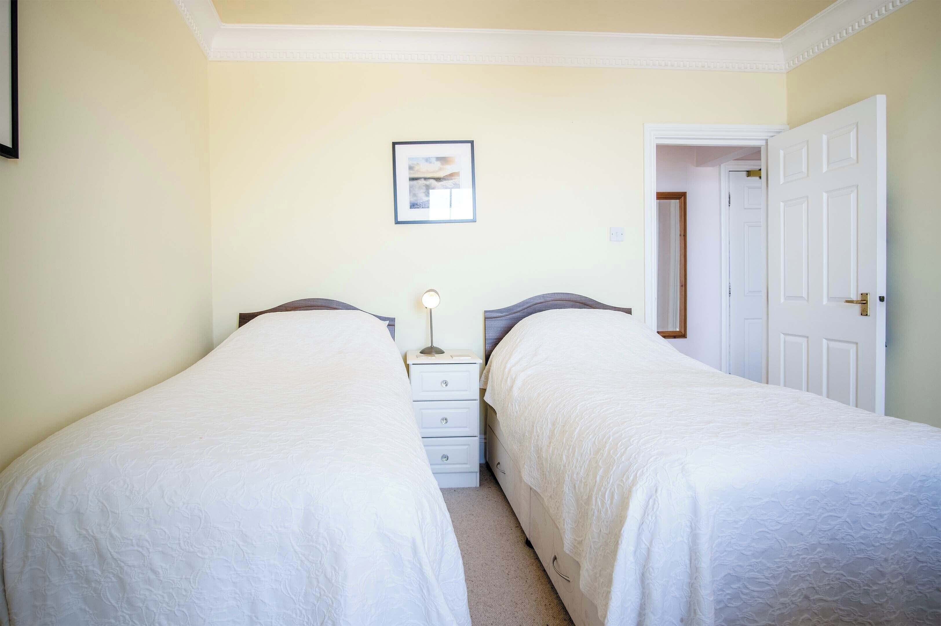 A bedroom with two single beds, a bedside table with a lamp, a framed picture on the wall, and a door leading to another room.