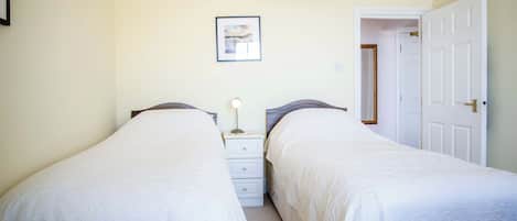 A bedroom with two single beds, a bedside table with a lamp, a framed picture on the wall, and a door leading to another room.