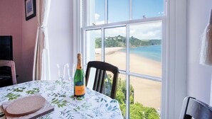 Une table dressée avec une bouteille de champagne, une assiette de pain et un verre, donnant sur une plage avec une vue dégagée sur la mer et les collines.
