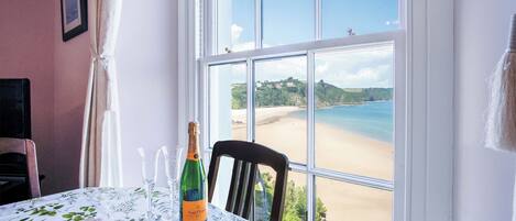 Une table dressée avec une bouteille de champagne, une assiette de pain et un verre, donnant sur une plage avec une vue dégagée sur la mer et les collines.