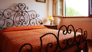 A bedroom with a bed, a nightstand, a lamp, a window, and a radiator. - Villa Amaranto Summar (Tuscany)