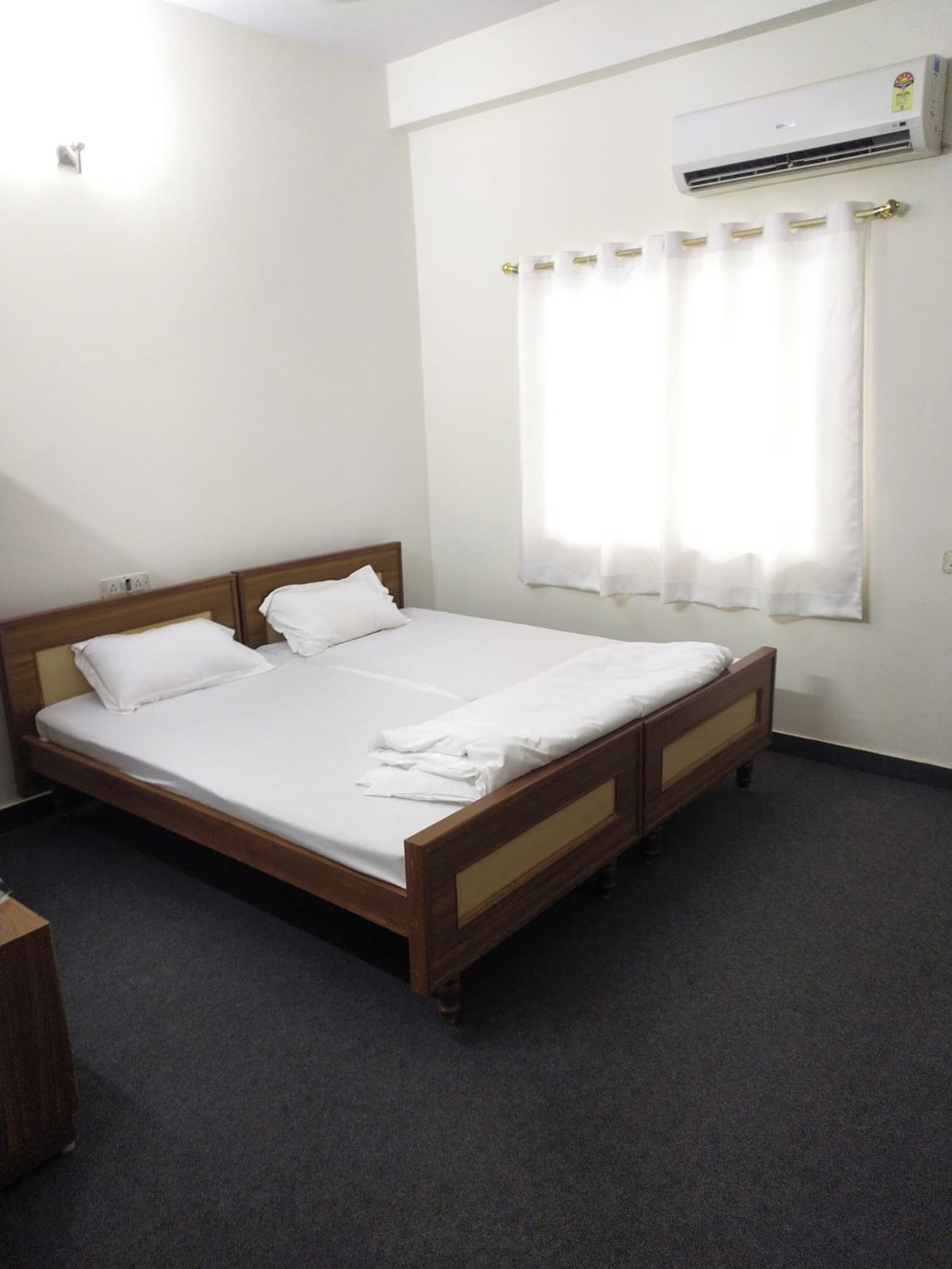 A single bed with wooden headboard and footboard, white bedding, a window with white curtains, and an air conditioning unit on the wall.