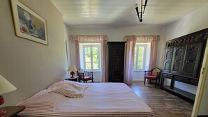 Une chambre à coucher avec un grand lit, deux fenêtres avec des rideaux, un plancher en bois, un bureau avec une lampe et une armoire.