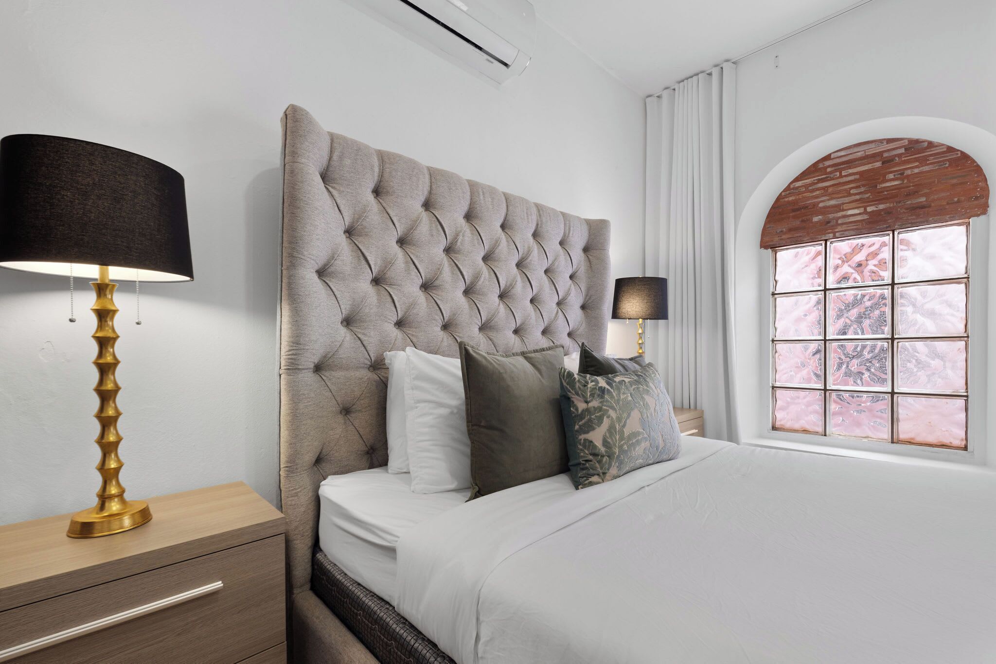 A bedroom with a tufted headboard, a wooden nightstand with a lamp, a bed with white bedding, and a window with a brick arch.