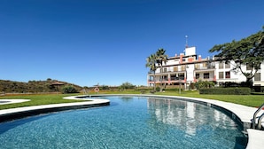 Une piscine d’un bleu éclatant, à bords incurvés, entourée d’une pelouse bien entretenue et de palmiers. Au fond, on aperçoit un grand bâtiment doté de nombreux balcons et d’un toit rouge.