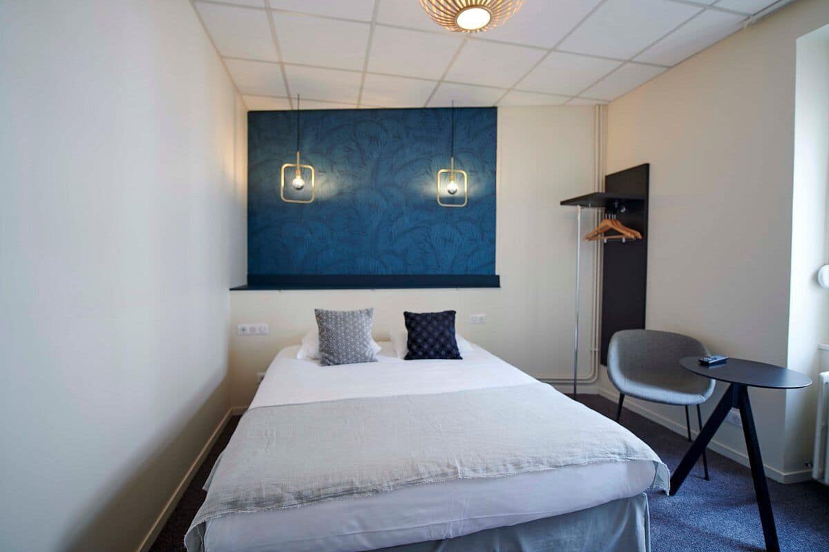 A hotel room with a large bed, two bedside lamps, a chair, a small table, and a coat rack.