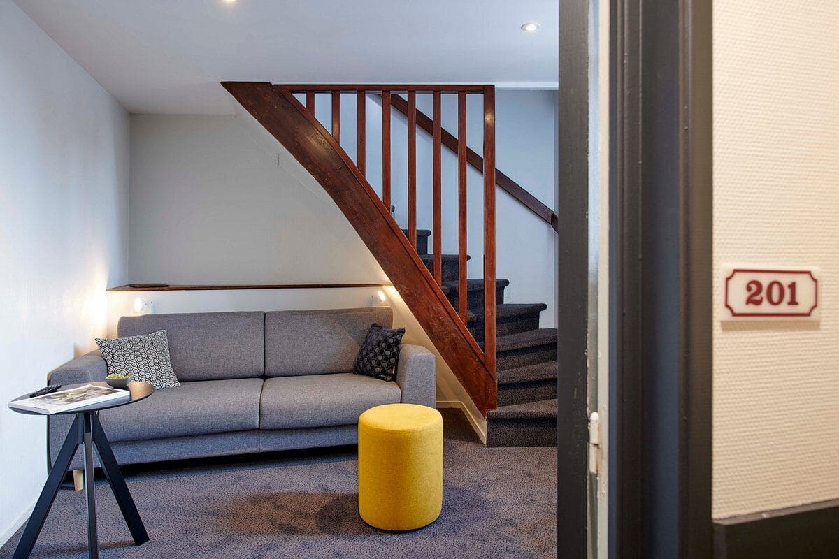 A modern living room with a grey sofa, a yellow ottoman, a small table, and a wooden staircase.