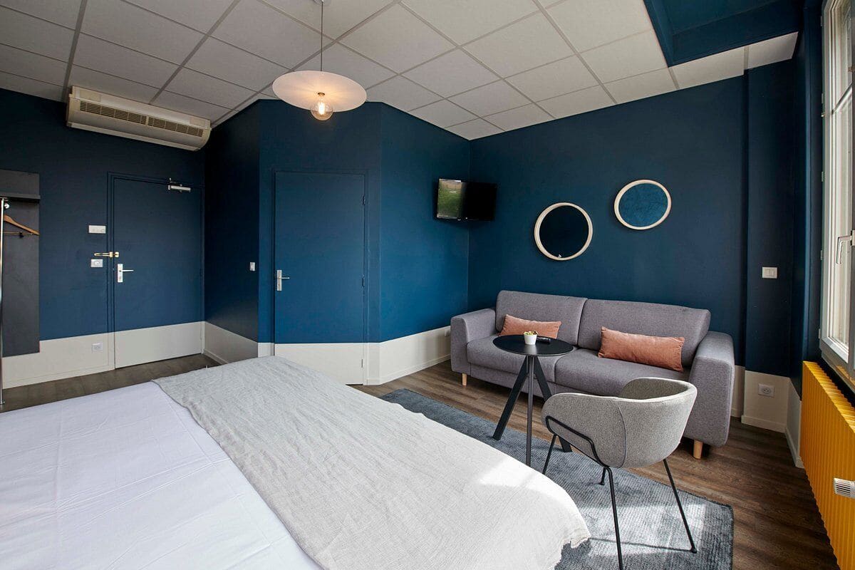 A modern hotel room with a large bed, a gray sofa, a small round table, and a TV mounted on the wall.