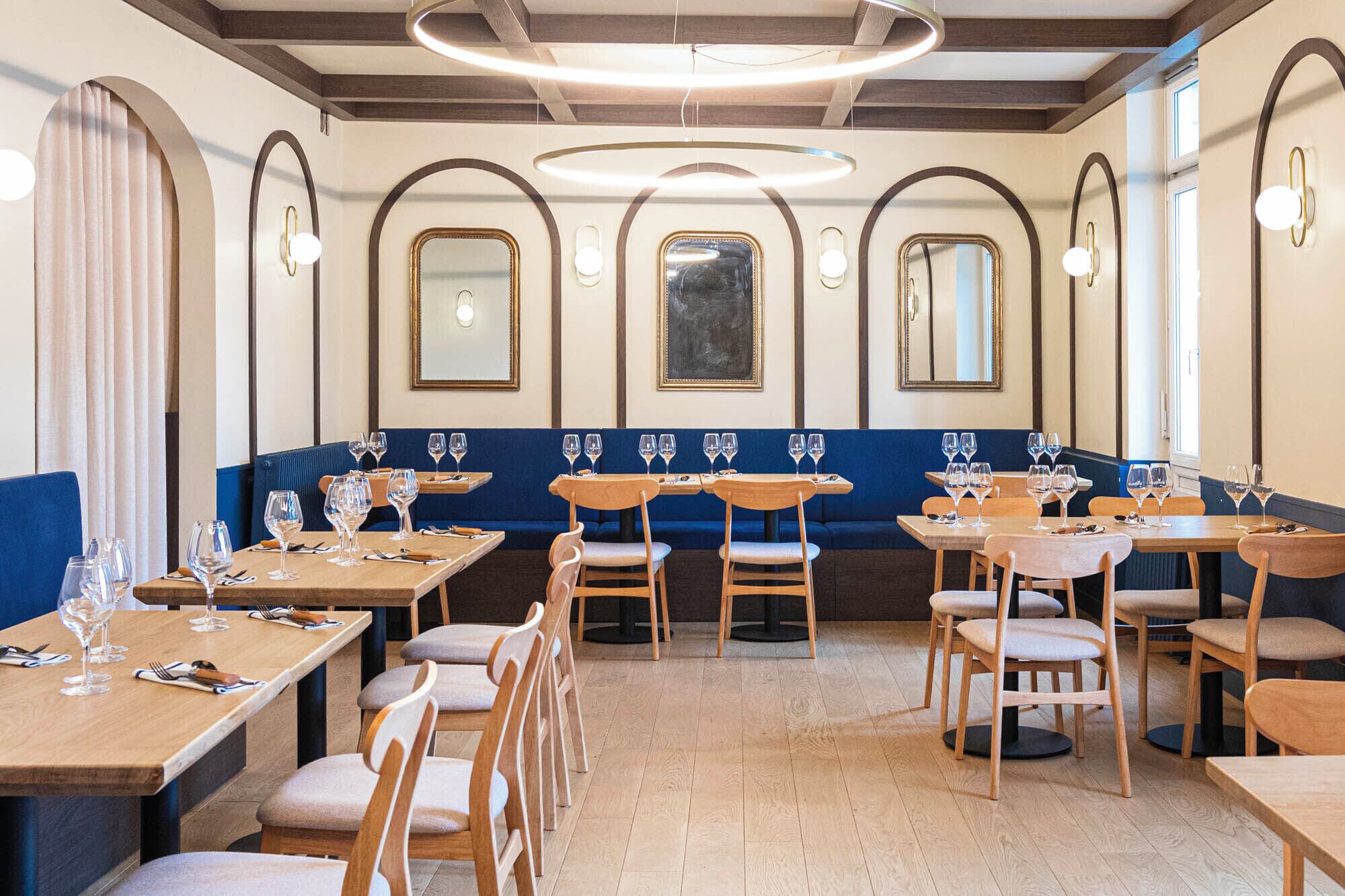 Un interior de restaurante con mesas y sillas de madera, asientos acolchados en azul y espejos decorativos en las paredes.