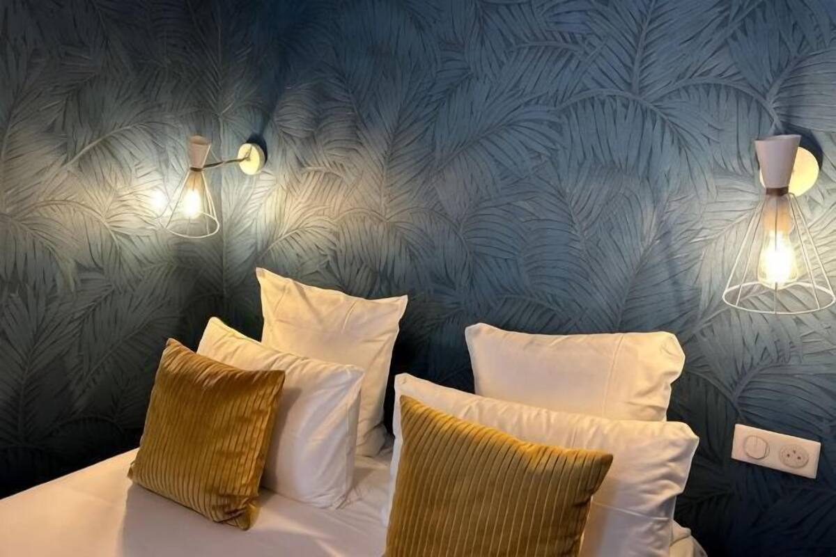 A room with a patterned wallpaper, a bed with white and gold pillows, and two wall-mounted light fixtures.