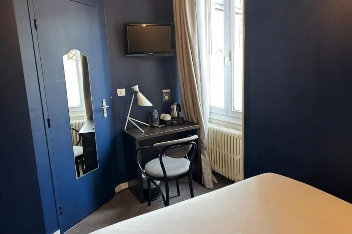 A hotel room with a bed, a desk with a chair, a TV mounted on the wall, and a window with curtains.
