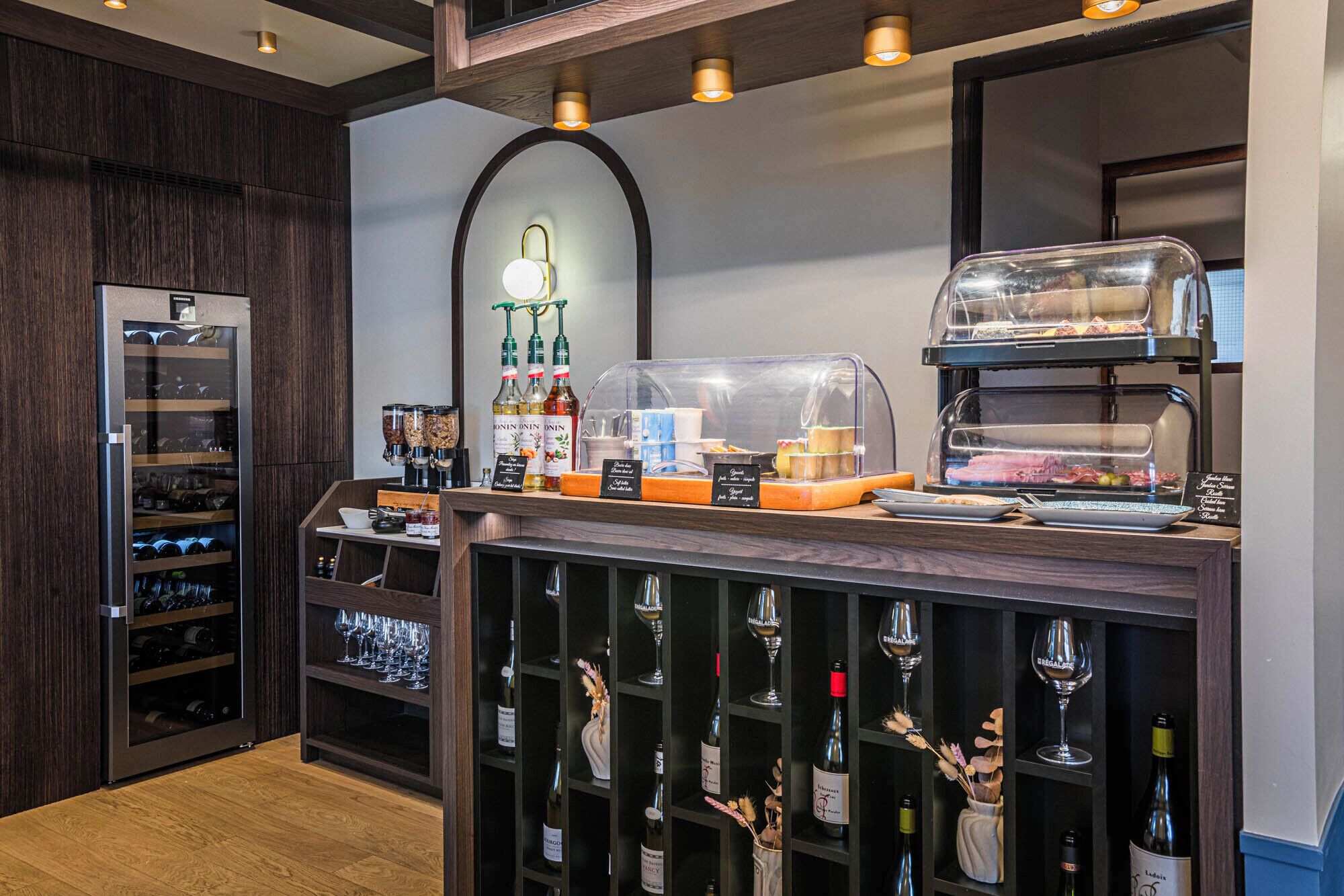 Un espace bar équipé d’un réfrigérateur à vin, d’un coin café et d’une vitrine réfrigérée où sont présentés des produits alimentaires.