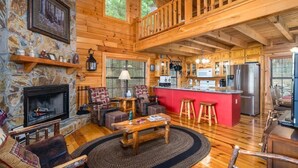 A cabin interior with a stone fireplace, wooden furniture, a kitchen area, and a balcony. - The Stargazers Perch (Blue Ridge)