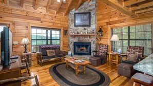 A log cabin interior with a stone fireplace, wooden furniture, and large windows. - The Stargazers Perch (Blue Ridge)