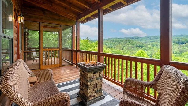 A wooden deck with wicker chairs and a stone fire pit, overlooking a lush green landscape. - The Stargazers Perch (Blue Ridge)