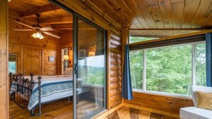 A log cabin interior with a bed, a sofa, a ceiling fan, and a large window offering a view of the outdoors. - The Stargazers Perch (Blue Ridge)