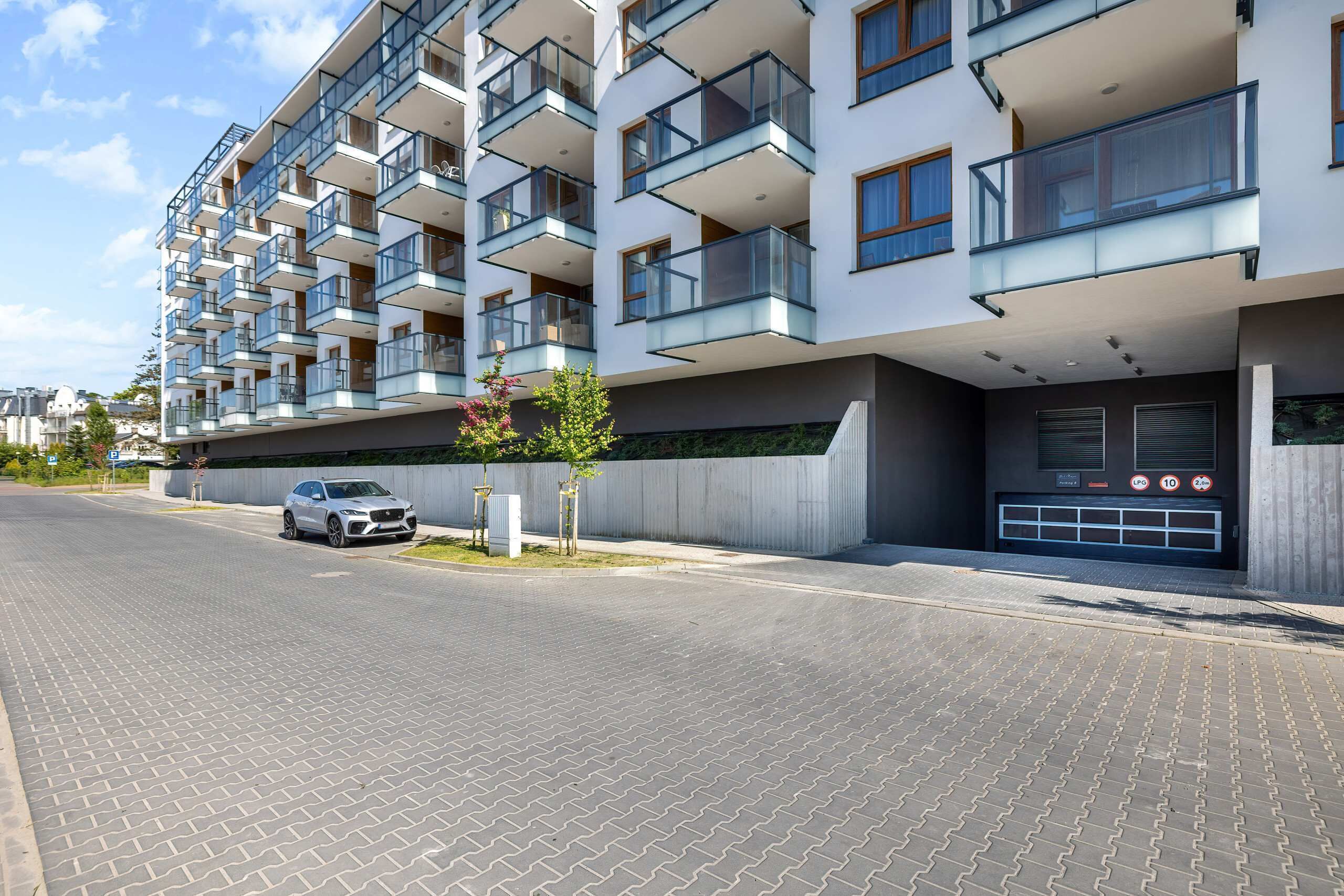 A multi-story residential building with balconies, a paved driveway, and a car parked in front.