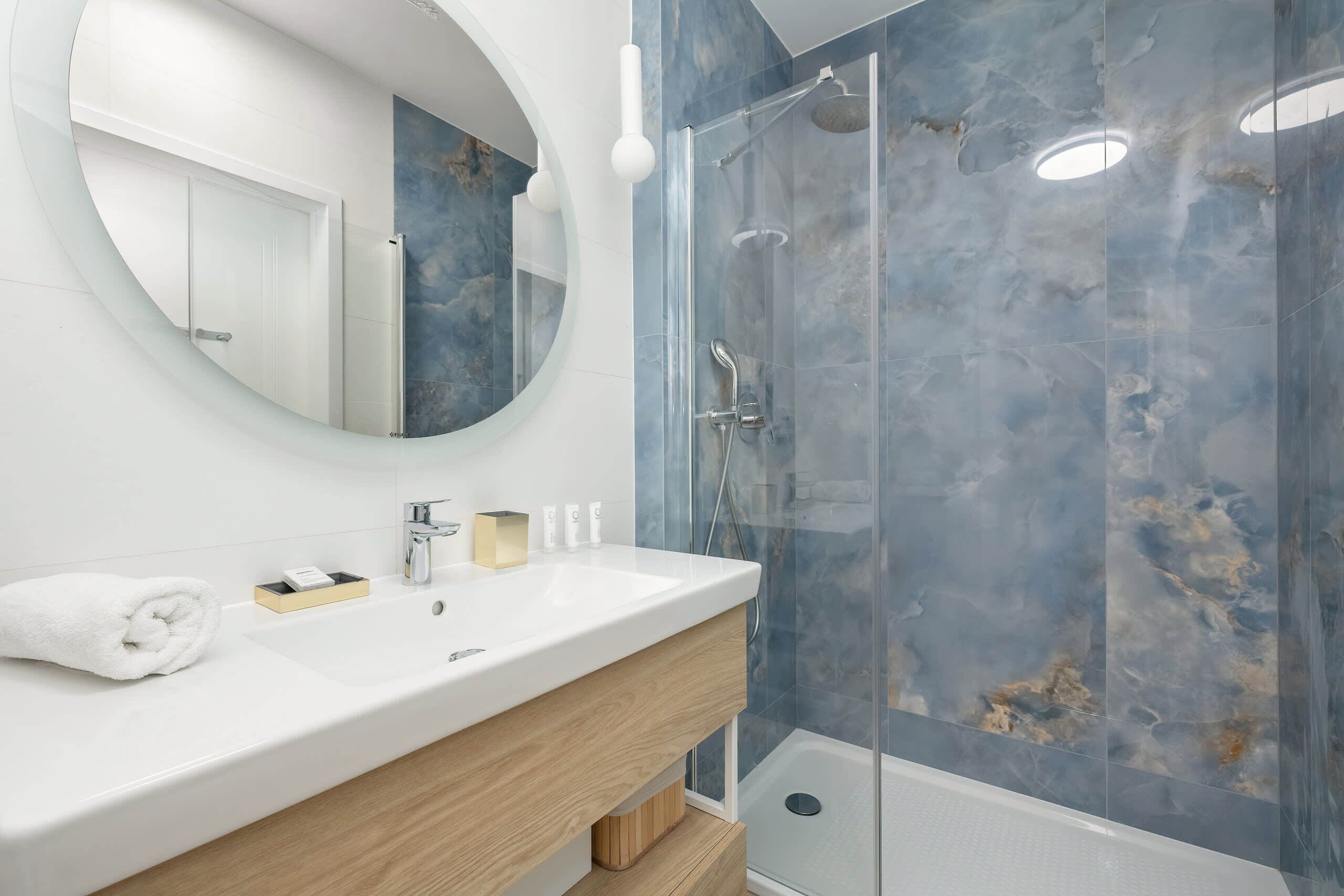 Un baño con un lavabo blanco, un espejo, una toalla, cabina de ducha de vidrio y pared de mármol.