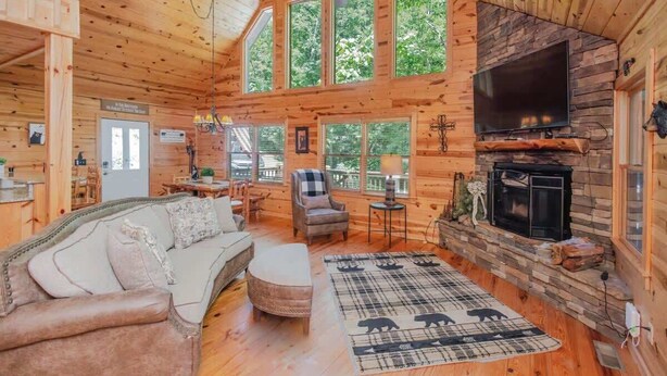 A log cabin interior with a fireplace, a TV mounted on the wall, a sofa, and a dining area. - Black Bear Lodge Summ (Blue Ridge)