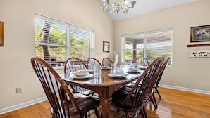 Comedor con una mesa de madera dispuesta para seis personas, sillas alrededor, ventanas amplias con persianas y una lámpara de araña.