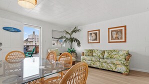 A modern living room with a glass dining table, wicker chairs, a floral sofa, and a large potted plant.