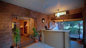 A reception desk with a man working on a laptop, a clock on the wall, and potted plants. - TreeHouse Rajbagh (Luni)