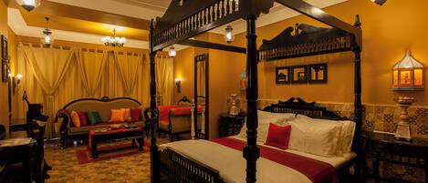 A four-poster bed with a red and white bedspread, a wooden bench, a sofa with colorful cushions, and a chandelier.