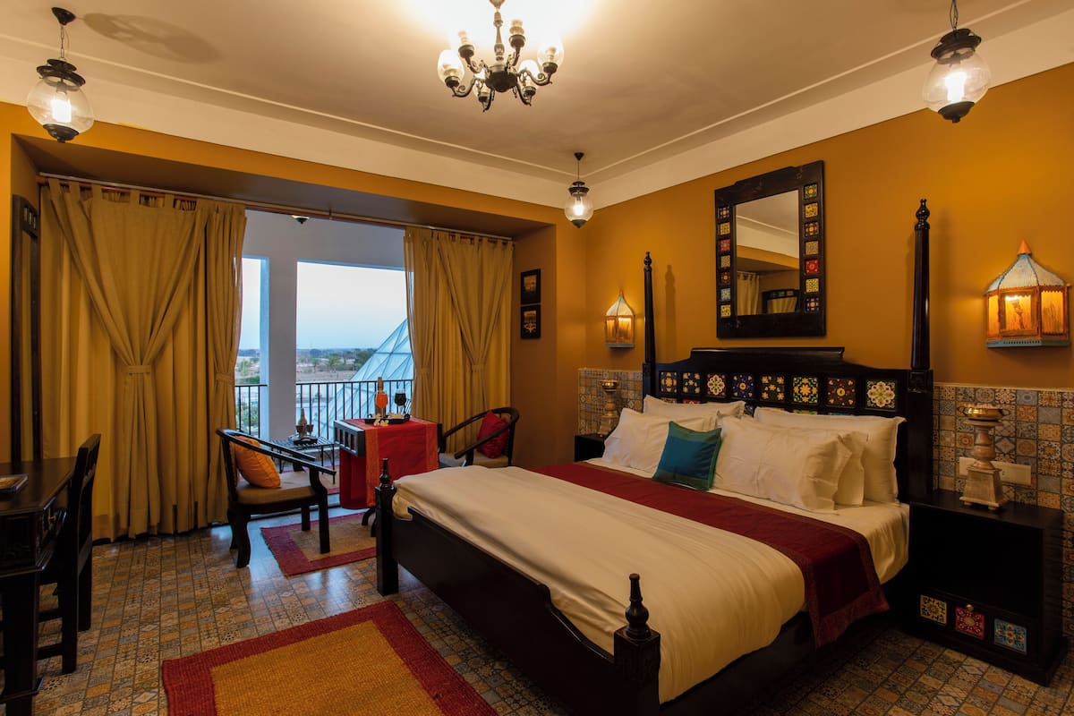Ein Hotelzimmer mit einem großen Bett, einem Schreibtisch und einem Sessel. Es gibt einen Balkon mit Vorhängen und Blick ins Freie.