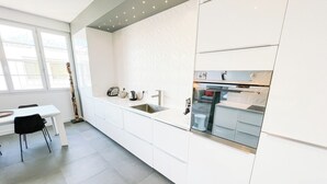 Une cuisine moderne avec des meubles blancs, un four encastré et une fenêtre donnant sur un quartier résidentiel.