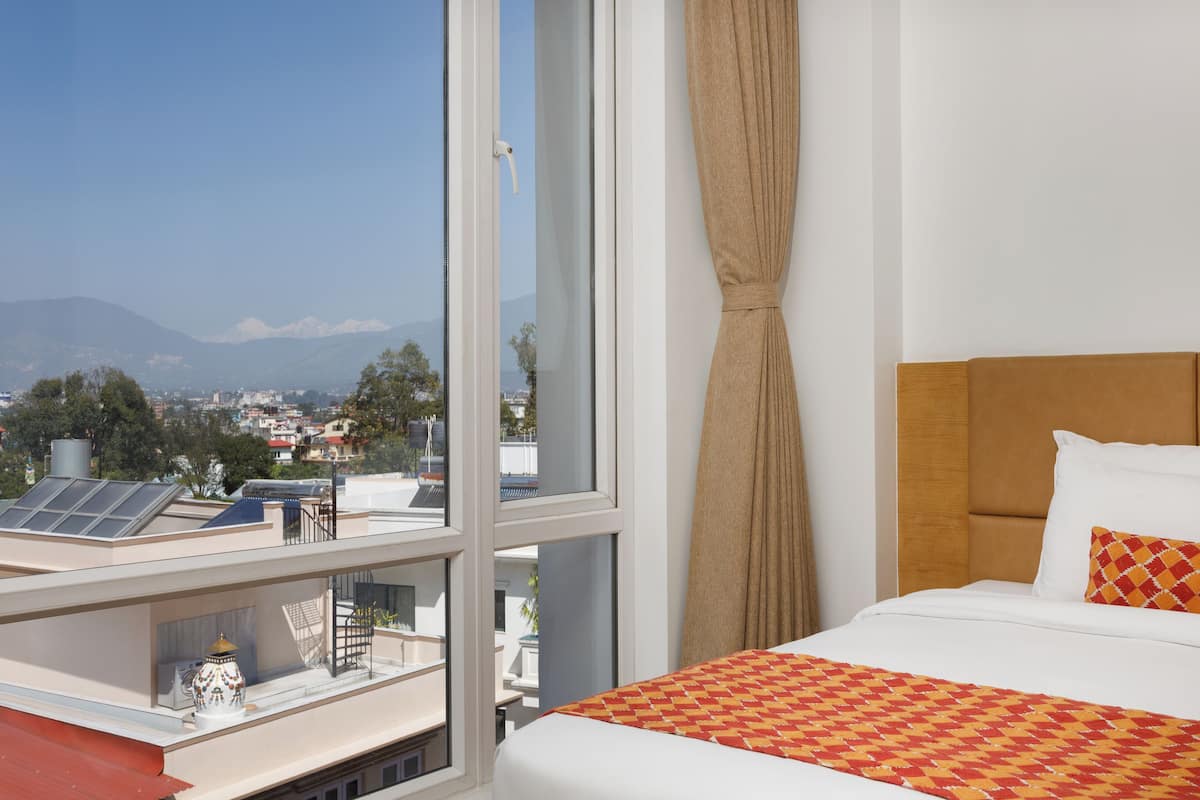 A hotel room with a large window, a bed with white bedding, and a view of a cityscape and mountains.