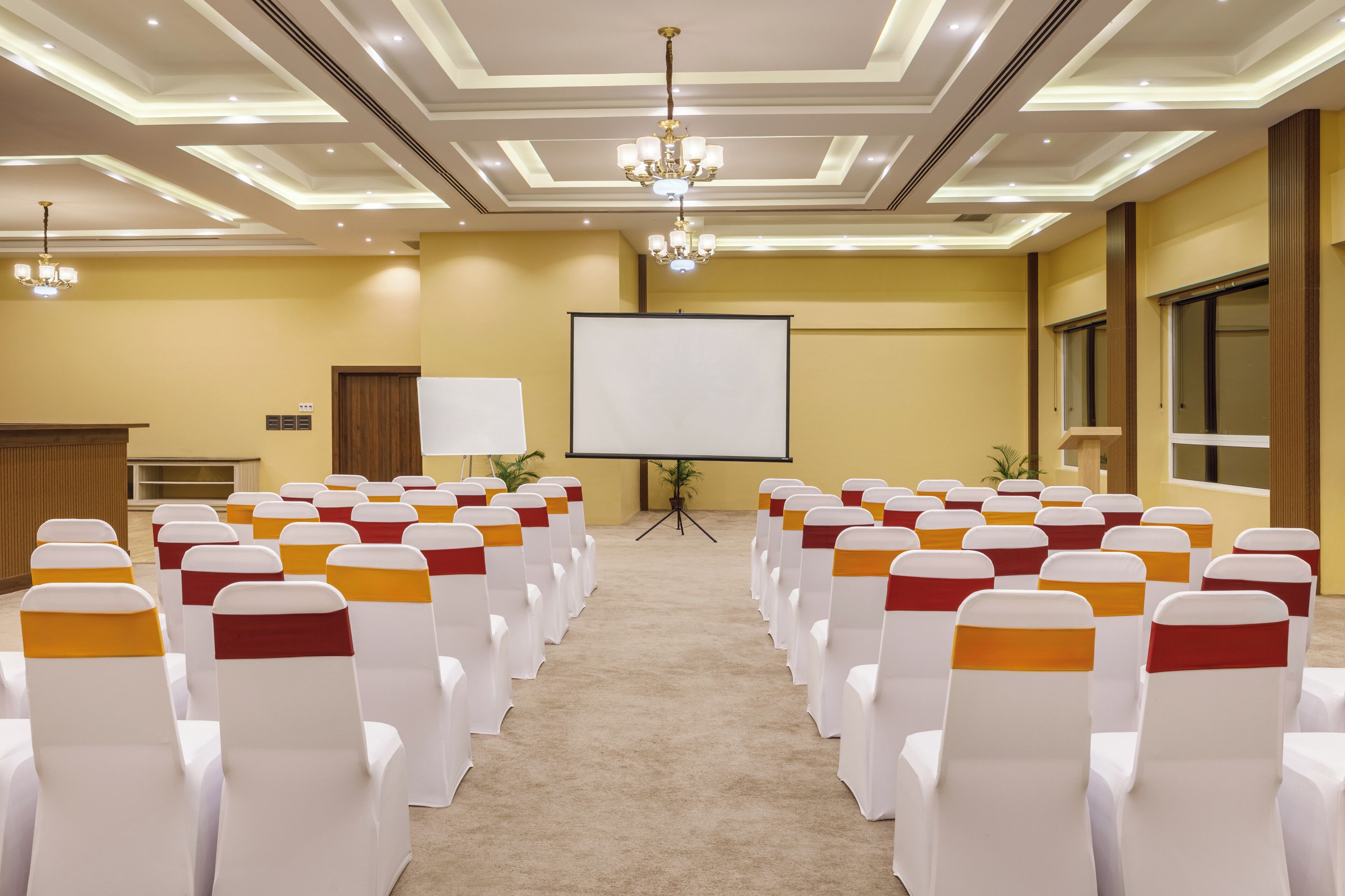 Une salle de conférence avec des rangées de chaises blanches ornées de rubans orange, un écran de projection et un pupitre.