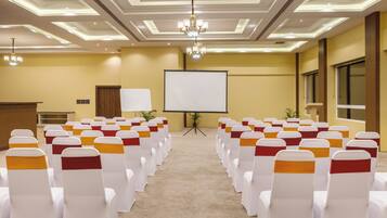 Une salle de conférence avec des rangées de chaises blanches ornées de rubans orange, un écran de projection et un pupitre.