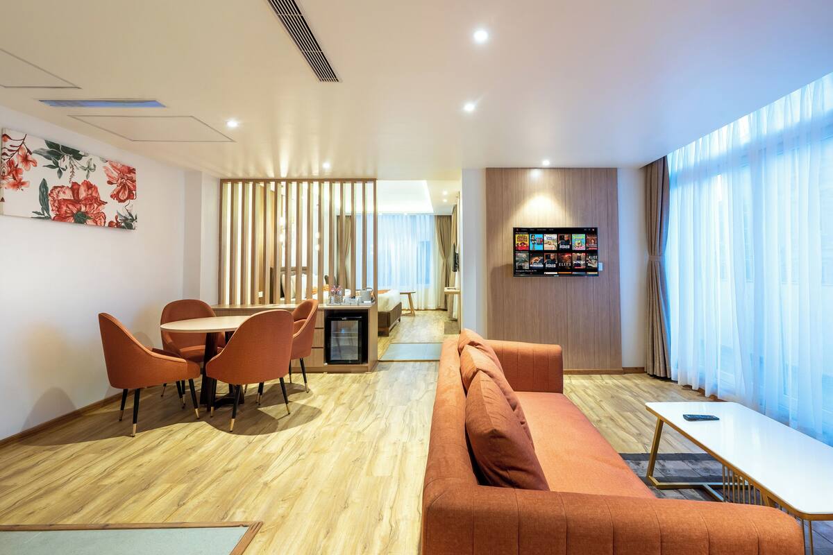 A modern hotel room with a wooden floor, orange sofa, dining table with chairs, a small kitchenette, and a large window with sheer curtains.