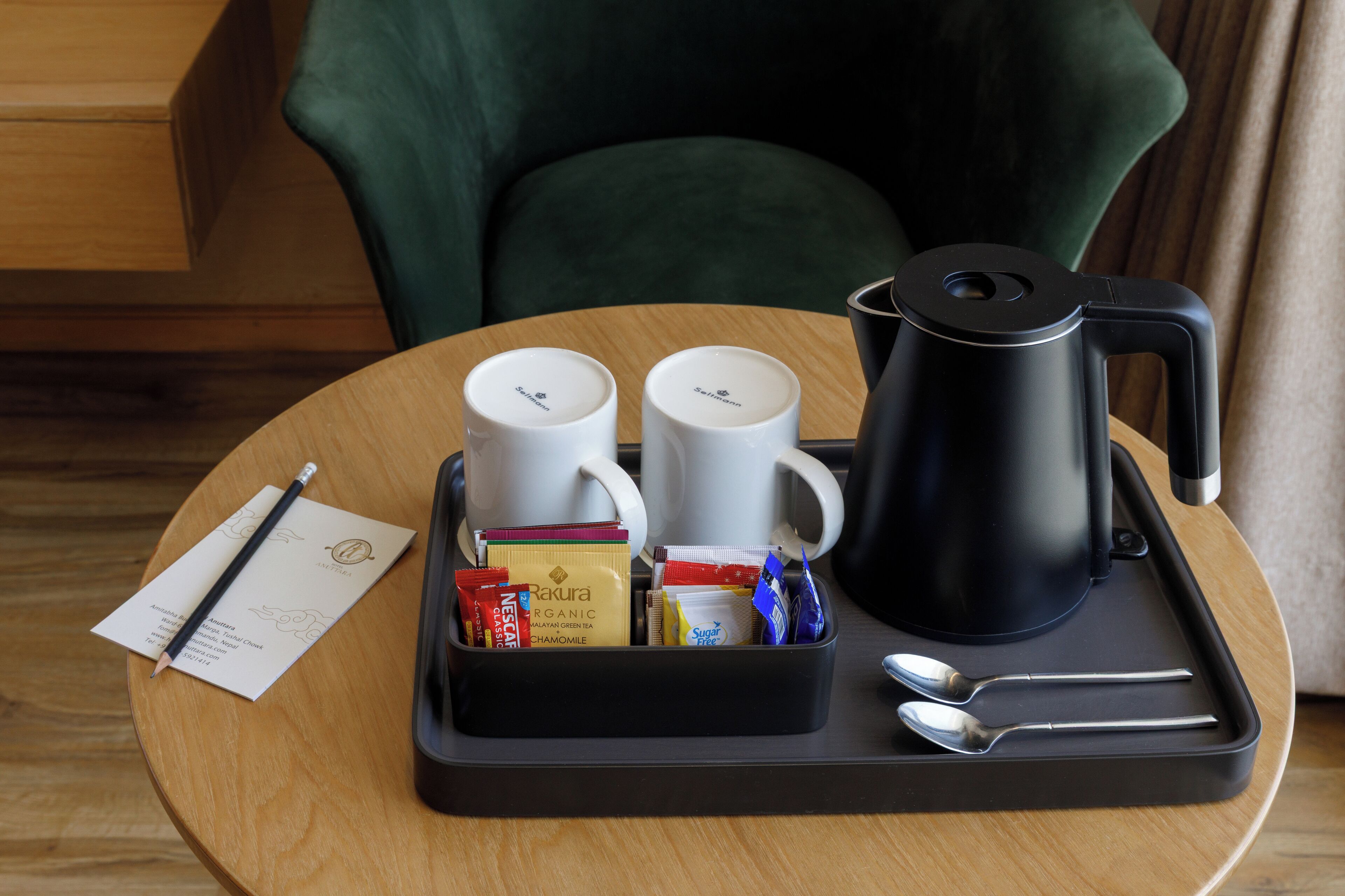 A tray with a black coffee maker, two white mugs, a spoon, a knife, and a small tray with snacks and a notebook on a wooden table.