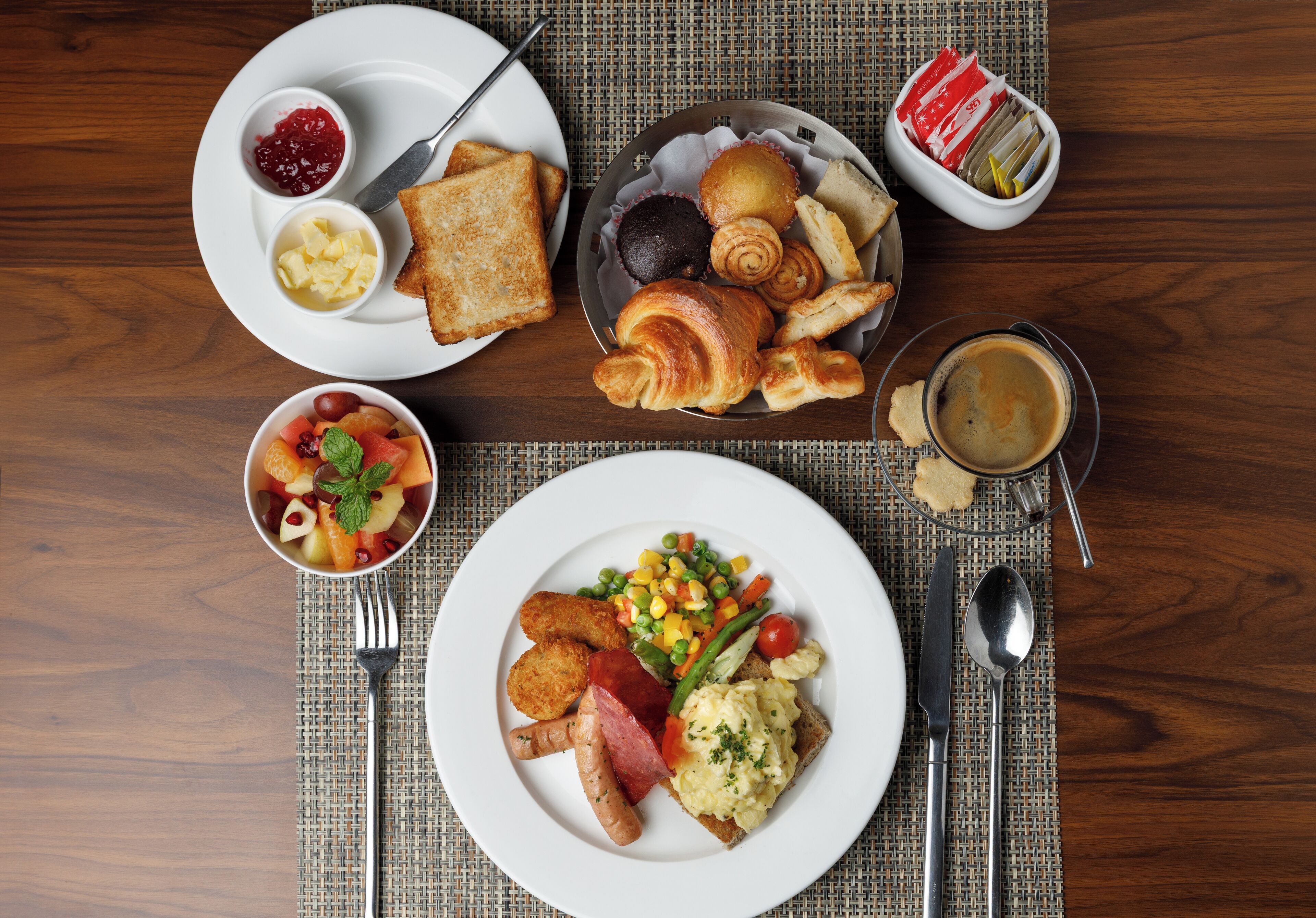 A breakfast setting with plates of toast, pastries, scrambled eggs, sausages, and a bowl of fruit salad.