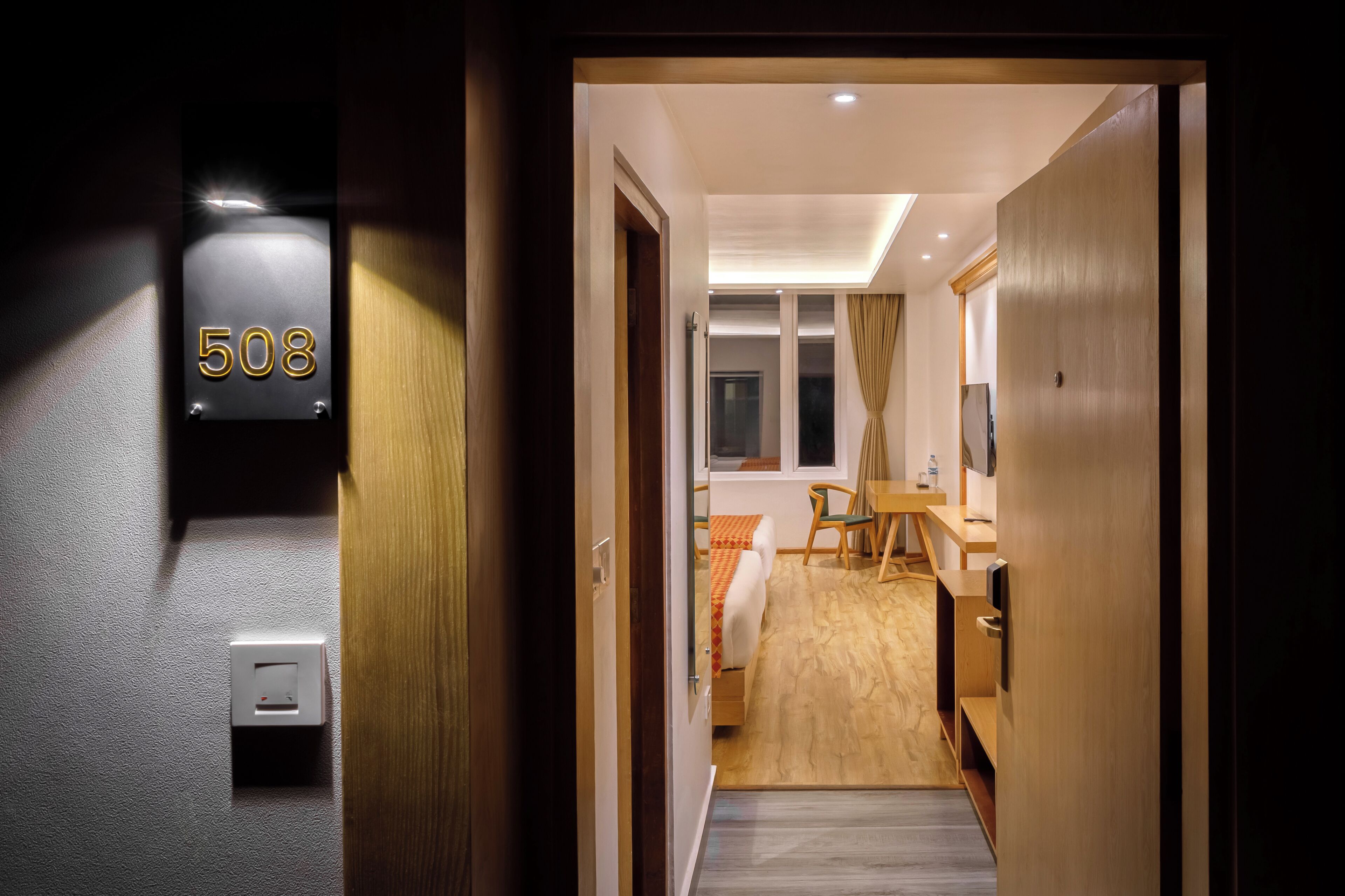 Une chambre d’hôtel avec un lit, un bureau et une chaise. Il y a un panneau avec un numéro sur le mur et un interrupteur.