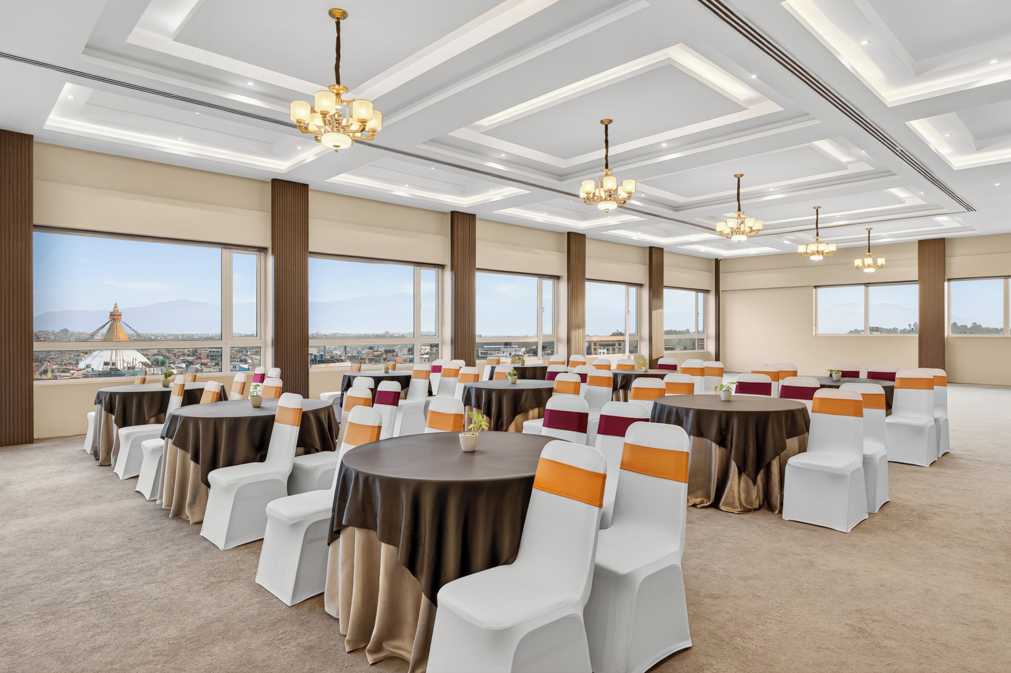 A conference room with round tables and white chairs, large windows with a city view, and modern chandeliers.