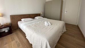 A neatly made bed with white linens and two rolled towels, a wooden nightstand with a lamp, and a wardrobe in a simple room. - PEL1900 (Cannes)