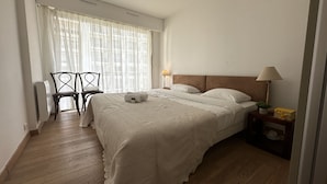 A bedroom with a bed, two bedside lamps, a chair, a small table with a lamp, and a window with sheer curtains. - PEL1900 (Cannes)