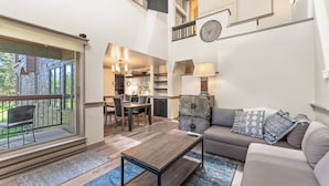 Een ruime woonkamer met een bank, salontafel en een eettafel. Achtergrond toont een keuken, en een groot raam zorgt voor natuurlijk licht.