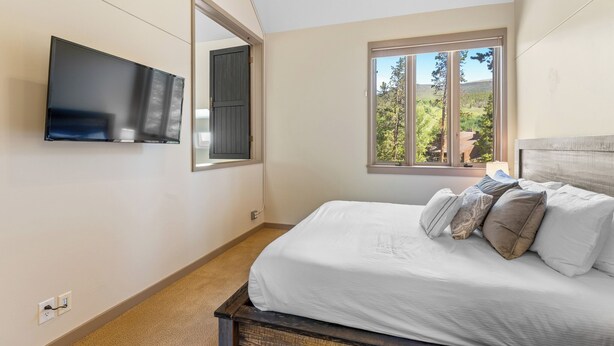 Een slaapkamer met een bed, een aan de muur bevestigde televisie, een raam met uitzicht op bomen, en een deur.