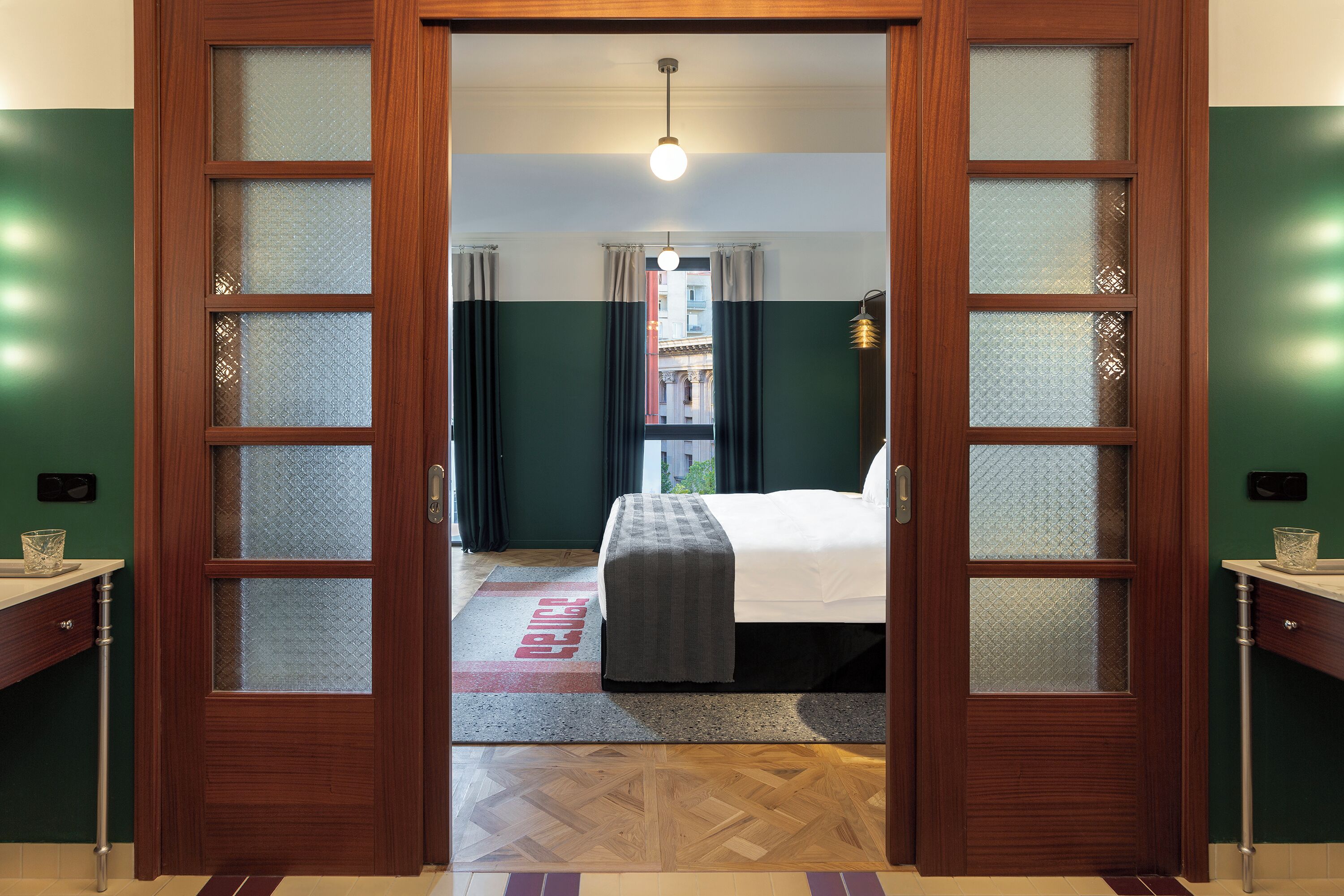 Una camera d'albergo con una porta in legno, un letto con coperta grigia, un comodino e vista sulla strada attraverso una finestra.